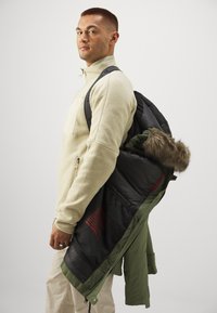 Pullover in pile beige chiaro per uomo con zip a un quarto, abbinato a una giacca nera e verde oliva con zip e cappuccio foderato in pelliccia.