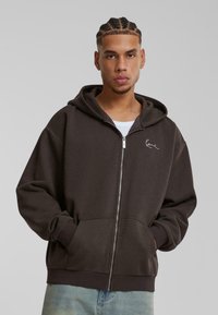 Sudadera con cremallera de color marrón oscuro, con un bolsillo frontal. Cuenta con capucha y un logo bordado en blanco en el pecho. Fabricada con una tela suave y texturizada.