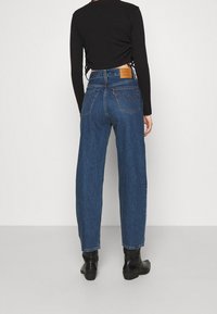 Jeans de cintura alta y corte suelto en denim azul oscuro. Presenta bolsillos traseros clásicos y un parche de cuero marrón en la cinturilla.