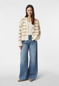Beżowo-kremowy kardigan w paski z dużymi guzikami, noszony na dopasowanym topie i szerokich, niebieskich dżinsach. Casualowa tkanina z prążkowaną fakturą.