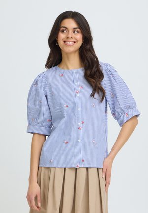 Ung kvinde med langt mørkt hår smiler iført en blå og hvid stribet bluse med pufærmer og små røde blomsterprint.