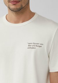 Crème-kleurige katoenen t-shirt met een ronde halslijn en korte mouwen, met in donkerbruin gedrukt tekst: "* kann Spuren von Bier und Burger enthalten."
