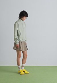 Une jeune personne se tient en regardant vers le bas, portant un pull-over gris clair, une jupe plissée beige, des chaussettes blanches rayées et des baskets jaunes sur un sol vert.