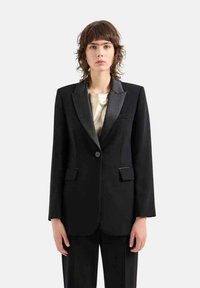 Blazer nero con rever in satin, chiusura a un bottone e due tasche frontali. Abbinato a una maglia metallica chiara sotto.
