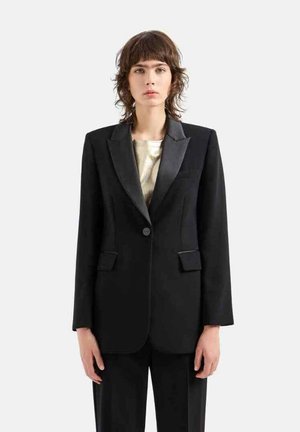 Blazer nero con rever in satin, chiusura a un bottone e due tasche frontali. Abbinato a una maglia metallica chiara sotto.