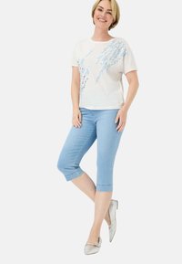 Kurzärmeliges weißes T-Shirt mit blauem Blumenmuster, kombiniert mit hellblauen Caprihosen und silbernen Ballerinas.