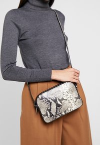 Osoba v šedém roláku a hnědých kalhotách drží crossbody tašku s černobílým hadím vzorem.