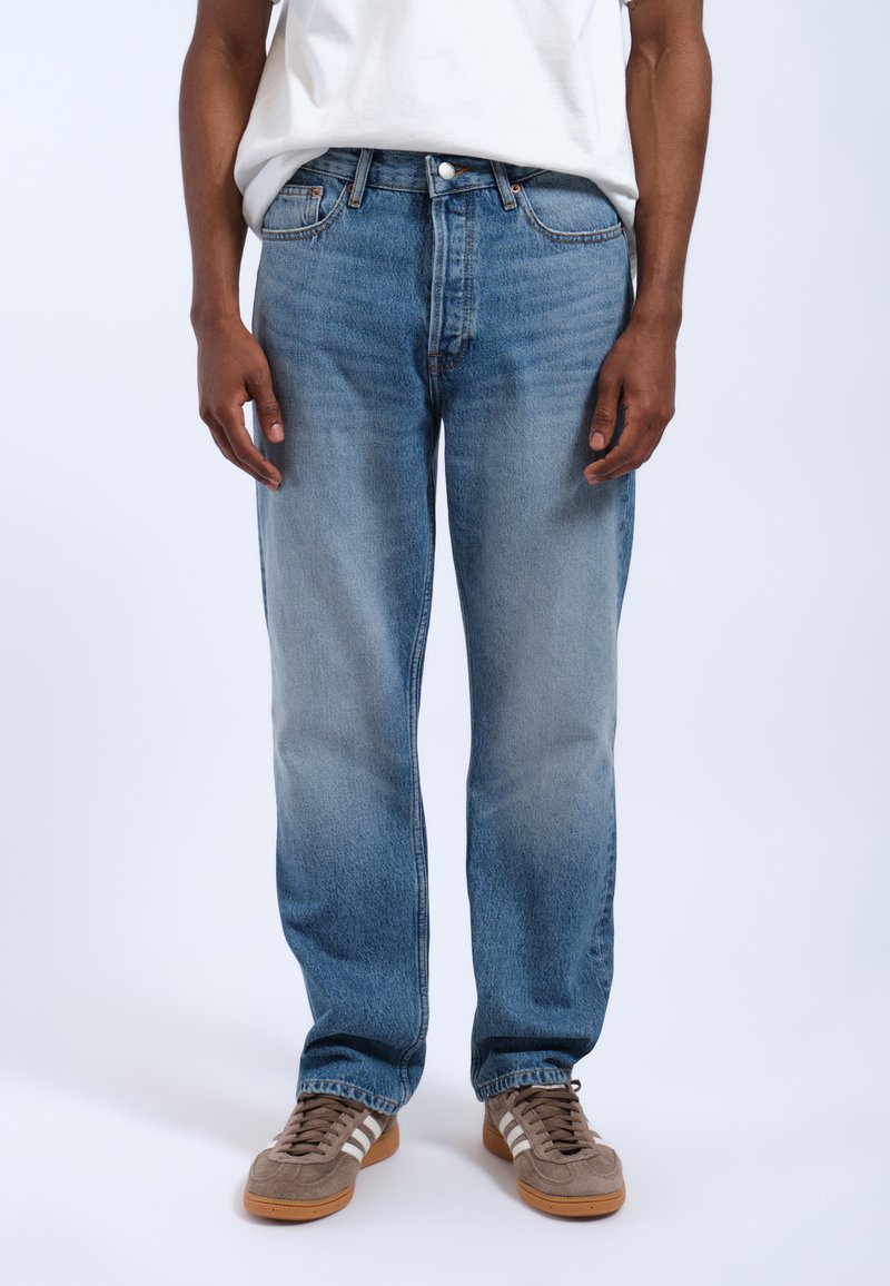 Jeans in denim blu chiaro con taglio dritto, dotati di cinque tasche, passanti per cintura e una finitura sfumata. Abbinati a scarpe marroni.