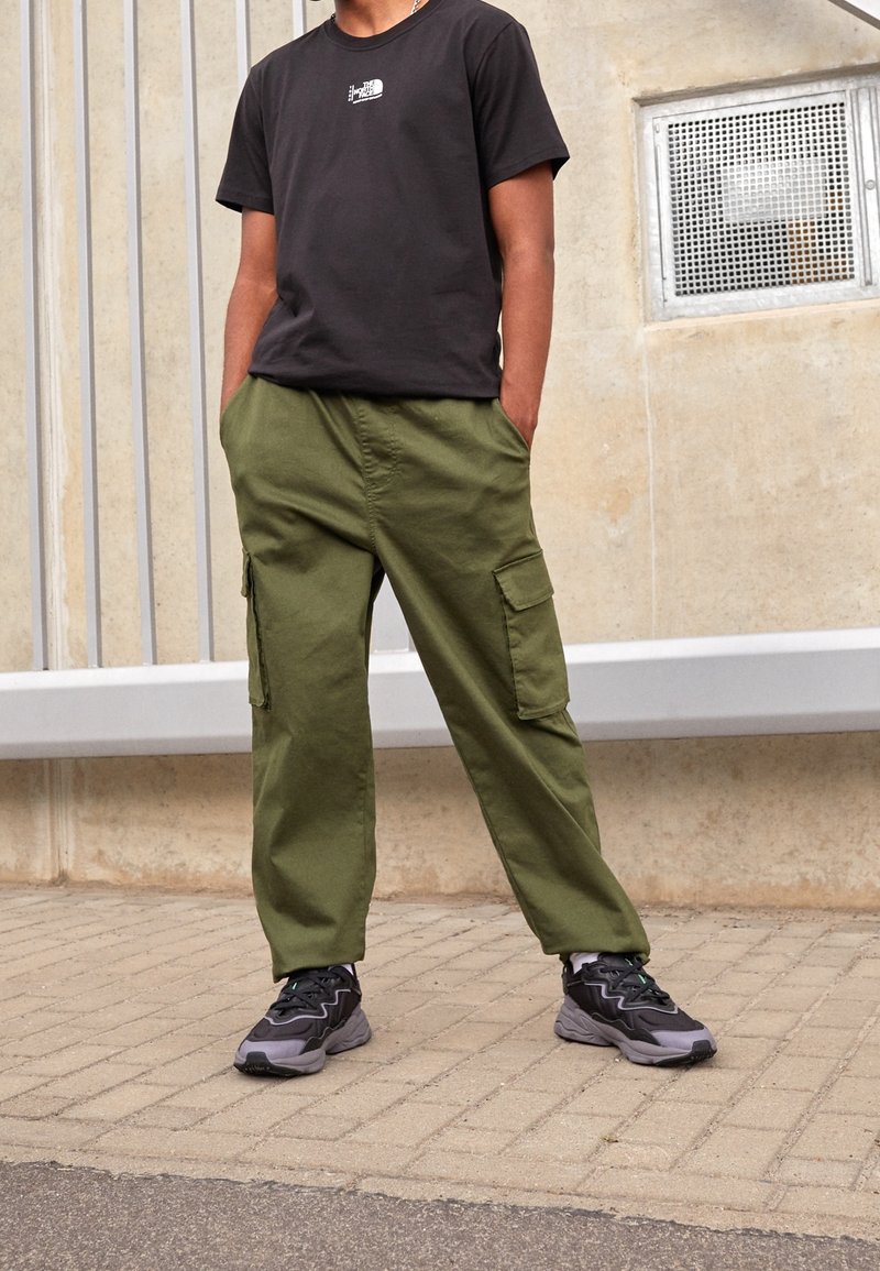 Camiseta negra de algodón con pequeño logo blanco, combinada con pantalones cargo verde oliva de corte holgado con bolsillos laterales y zapatillas negras con detalles.