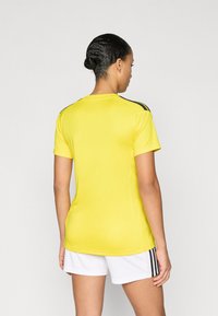 Κίτρινη αθλητική μπλούζα με κοντά μανίκια, στρογγυλή λαιμόκοψη και μαύρες ρίγες στους ώμους. Μαλακό, υφασμάτινο υλικό με υφή.