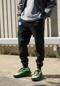 Sorte joggingbukser med sidelommer, prydet med et blåt logo og et rødt Nike-swoosh. Grønne sneakers med en struktureret overflade og markant dæmpning.