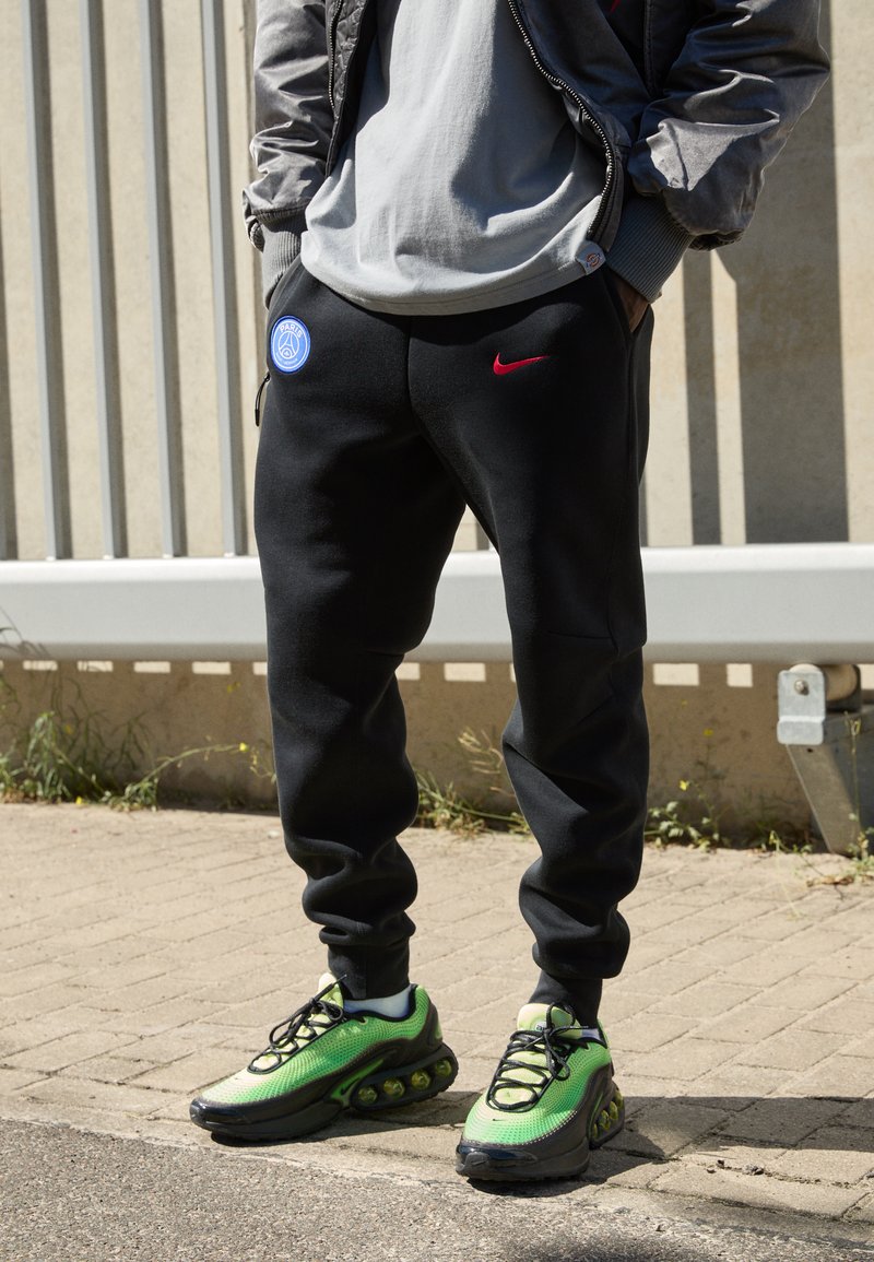 Sorte joggingbukser med sidelommer, prydet med et blåt logo og et rødt Nike-swoosh. Grønne sneakers med en struktureret overflade og markant dæmpning.