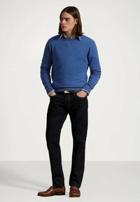 Homem com cabelo à altura dos ombros, vestindo um suéter azul, camisa riscada, jeans escuros, cinto castanho e sapatos castanhos, com as mãos nos bolsos, olhando para a direita.