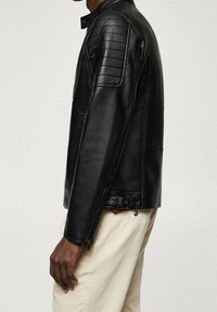 Giacca da moto in pelle nera con pannelli trapuntati sulle spalle, colletto alzato e cerniere laterali, abbinata a pantaloni in corduroy color crema.