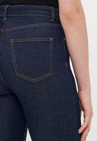 Close-up of a person’s lower back and hand wearing dark blue denim jeans with visible orange stitching and a back pocket.