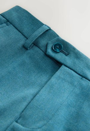 Gros plan sur un pantalon en tissu bleu canard avec une ceinture, passant de ceinture, bouton et poche avant sur fond blanc.