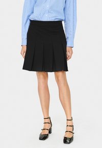 Jupe plissée noire avec une taille ajustée, associée à une chemise rayée bleu clair. La jupe tombe au-dessus du genou, avec une texture lisse.