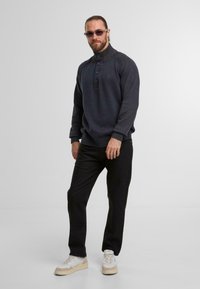 Maglione a maglia grigio scuro con colletto alto, tre bottoni al collo, polsini a coste, abbinato a pantaloni neri e scarpe da ginnastica chiare.