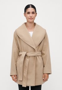 Kvinne med mørkt hår samlet bak, iført en beige belteullkåpe over en hvit topp, stående mot en lys plain bakgrunn.