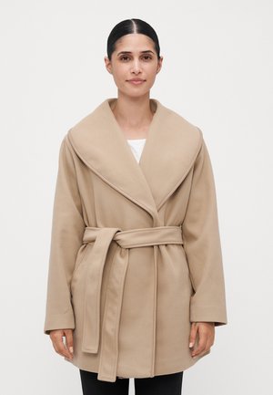 Donna con capelli scuri legati indietro, che indossa un cappotto di lana beige con cintura sopra una maglietta bianca, in piedi contro uno sfondo chiaro e semplice.