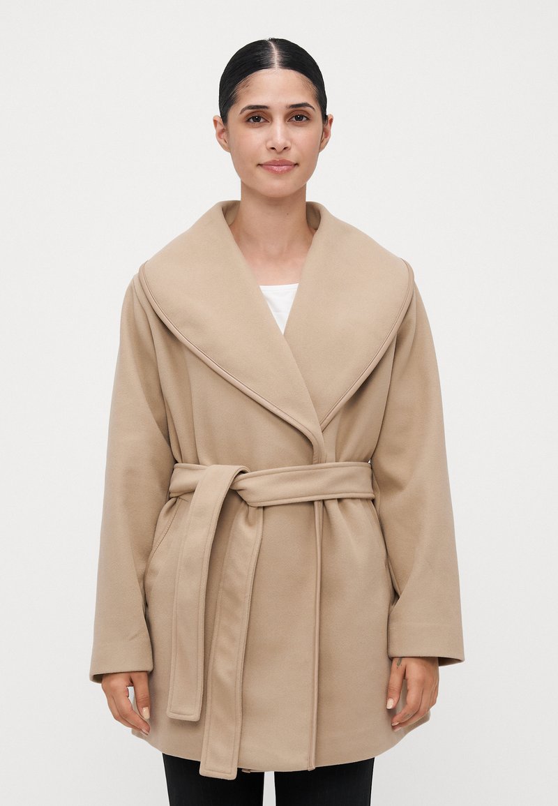 Kvinne med mørkt hår samlet bak, iført en beige belteullkåpe over en hvit topp, stående mot en lys plain bakgrunn.