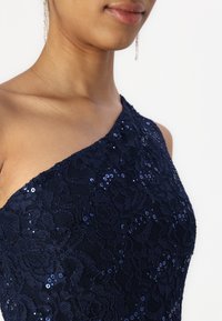 Robe en dentelle marine avec un décolleté asymétrique, ornée de sequins. Présente un motif floral texturé et une silhouette ajustée.