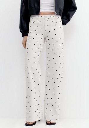 Fehér, széles szárú nadrág fekete pöttyökkel, egy személy viseli, aki rövid fehér felsőt és fekete dzsekit hord.