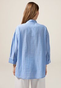 Camicia a righe blu realizzata in tessuto leggero. Presenta maniche lunghe con polsini a bottoni e una vestibilità comoda, con un orlo curvo sul retro.
