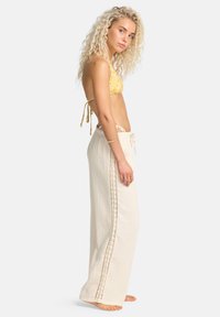 Ελαφριά, μπεζ παντελόνια με φαρδιά πόδια και λεπτομέρειες δαντέλας κατά μήκος των πλευρών και ζώνη με κορδόνι, φορεμένα πάνω από κίτρινο μπικίνι με λουλούδια.