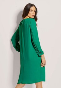 Grün Kleid mit langen Ärmeln, das über gefaltete Details und einen Knopfverschluss auf der Rückseite verfügt. Glattes Gewebe mit einem lockeren Schnitt.