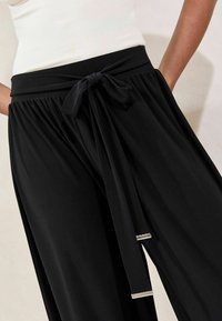 Schwarze weite Hosen aus weichem Stoff, mit einem gebundenen Taillengürtel und Seitentaschen, sowie einem silbernen Akzent am Hosenbein.