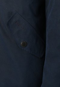 Sötétkék viaszolt pamut dzseki, amelyen egy dombornyomott "Barbour" logós fedélzseb és egy sötét fém snap gomb található.