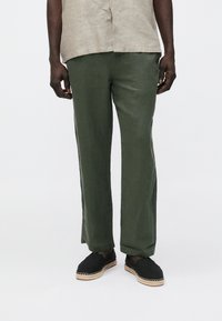 Olivgrüne Leinenhose mit entspanntem Schnitt, seitlichen Schlitzen und kombiniert mit schwarzen Espadrilles mit gewebten Juteverzierungen.