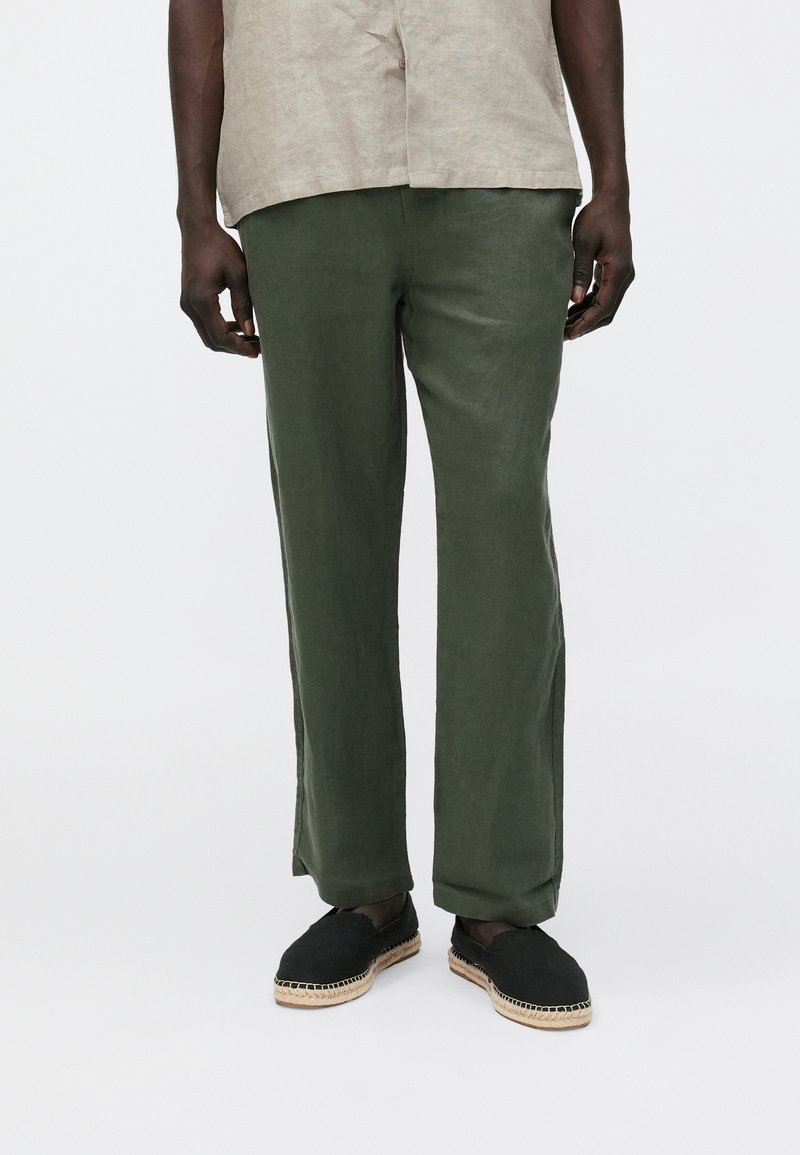 Olivgrüne Leinenhose mit entspanntem Schnitt, seitlichen Schlitzen und kombiniert mit schwarzen Espadrilles mit gewebten Juteverzierungen.