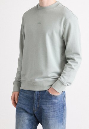 Mann trägt hellgraues Sweatshirt mit Rundhalsausschnitt und "BOSS"-Logo sowie blaue Jeans, eine Hand in der Tasche, vor schlicht weißem Hintergrund.