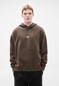 Rudas džemperis su gobtuvu, turintis tekstūrinę apdailą, priekinę kišenę ir mažą baltą stačiakampį logotipą. Turi raištelį ant gobtuvo ir laisvą siluetą.