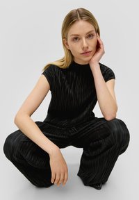 Zwarte geplooide jumpsuit met hoge kraag, korte mouwen en losse broekspijpen. De stof heeft een glanzende textuur en een verticale ribbelpatroon.