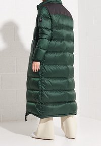 Cappotto imbottito lungo verde con pannello nero sulla spalla, caratterizzato da cuciture orizzontali, colletto alto e chiusura con zip. Tessuto liscio.