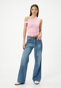 Top ajustado rosa de un solo hombro con textura fruncida, combinado con jeans de mezclilla claros de pierna ancha. La modelo está de pie con una mano en la cadera.