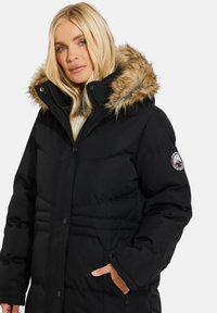 Sort pufferjakke med hætte trimmet med pels, designet med lynlås og trykknap lukninger, quiltet tekstur og et logo patch på ærmet.