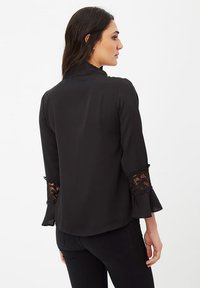 Blusa negra con acentos de encaje transparente en las mangas. Tela suave con un corte holgado y puños acampanados. La vista trasera resalta los detalles del diseño.