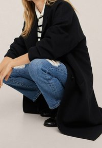 Manteau noir surdimensionné avec de grands revers, porté sur un haut rayé blanc et noir. Jean bleu déchiré et chaussures noires brillantes.