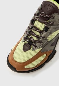 Limezöld hálós sneaker barna bőráccentelekkel, szürke szövetpanelek és fekete gumitalp. Sötétbarna fűzőkkel és megerősített orrkappal rendelkezik.