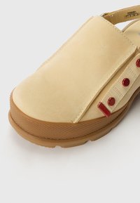 Geel leren instappers met een ronde neus, een gestructureerde rubberen zool en rode accentknopen aan de zijkant.