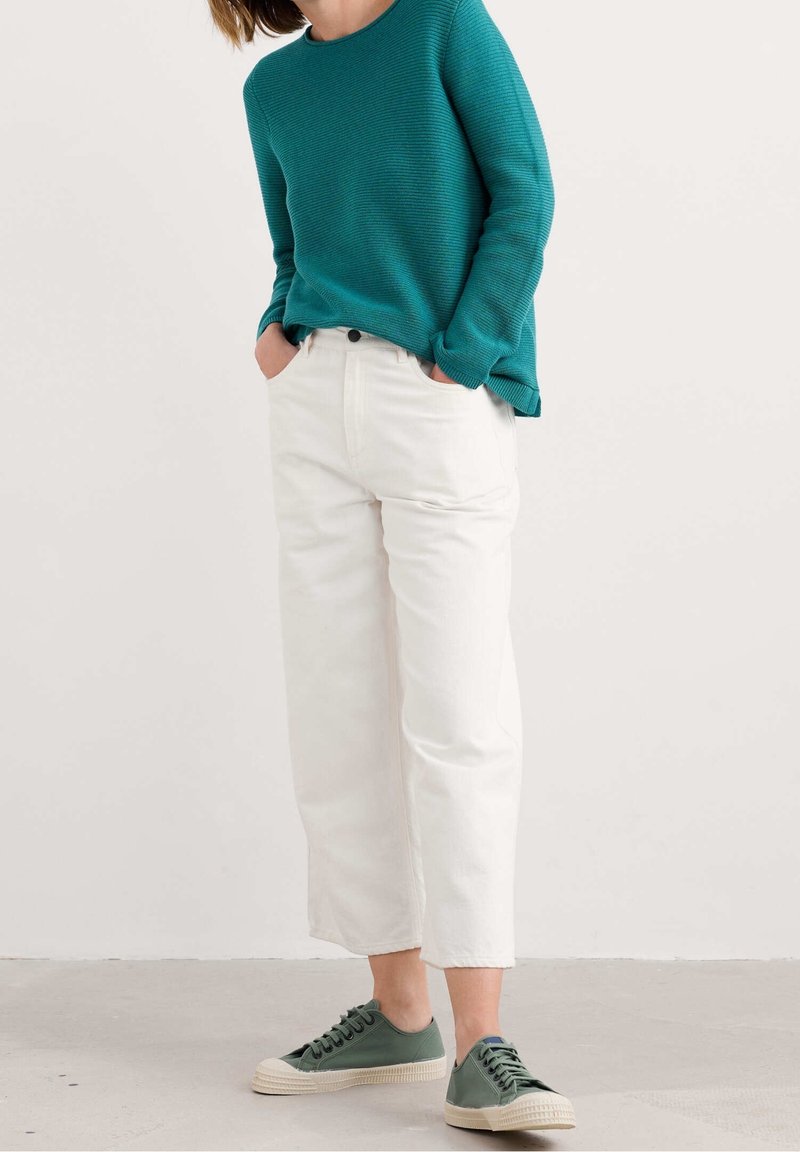 Personne portant un pull à manches longues turquoise, un pantalon blanc court taille haute, et des baskets basses vertes, debout les mains dans les poches.