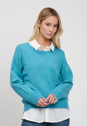 Mujer con cabello largo rubio lleva un suéter azul sobre una camisa blanca con cuello, manos entrelazadas al frente, fondo gris neutro.
