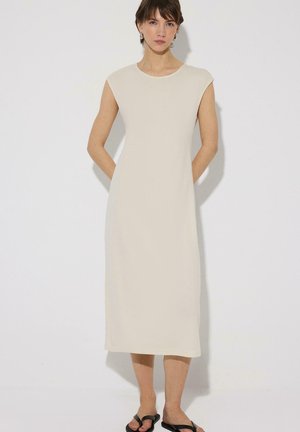 Femme debout contre un mur blanc, portant une robe midi beige sans manches et des sandales noires, les mains dans le dos, expression neutre.