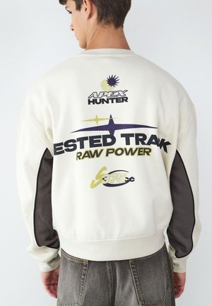 Junger Mann trägt ein weißes Sweatshirt mit schwarzem und gelbem grafischem Text und schwarzen Patches an den Ärmeln, von hinten gesehen.