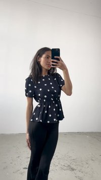 Blusa preta com bolinhas, com design peplum, mangas curtas e volumosas, e cintura ajustada, combinada com calças pretas sobre um fundo neutro.