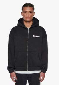 Sort hoodie med lynlås, der har en forlommer og broderet hvidt logo. Lavet af blødt stof, med en hyggelig hætte og elastiske manchetter.